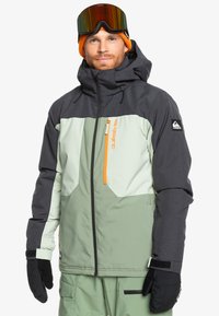 Chaqueta aislante para hombre con un diseño de bloques de color en gris, verde menta y verde claro, que presenta un cierre naranja y guantes negros.