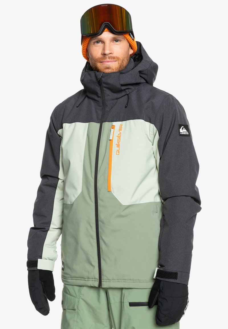 Chaqueta aislante para hombre con un diseño de bloques de color en gris, verde menta y verde claro, que presenta un cierre naranja y guantes negros.