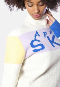 Farbblockierter Rollkragenpullover in Creme, Lavendel, Gelb und Blau. Verfügt über eine gerippte Textur und gestrickte Buchstaben auf der Brust.