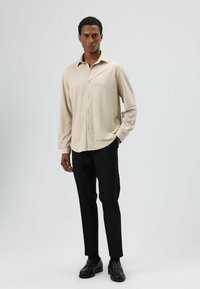 Beige lange mouwen overhemd met knopen en een gladde textuur, gedragen met zwarte, op maat gemaakte broek en zwarte loafers, staand tegen een lichtgrijze achtergrond.