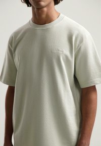 Tricou verde deschis dintr-un material moale, cu guler rotund ribbufat și mâneci scurte. Prezintă un detaliu subtil cu logo brodat pe piept.