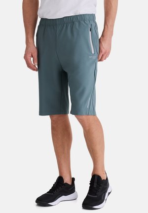 Mann trägt türkisblaue knielange Sportshorts mit seitlichen Reißverschlusstaschen und schwarze Sportschuhe vor weißem Hintergrund.