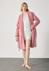 En rosa terry-kåpe med teksturert overflate, med to frontlommer, brukt over hvite shorts og en kortermet topp med blondebesetning.