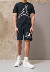 T-shirt noir avec un motif Jordan, associé à un short noir avec un logo blanc. Complété par des baskets montantes blanches et des chaussettes blanches.