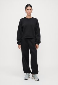Černá mikina s kulatým výstřihem a spadlými rameny, v kombinaci s odpovídajícími černými tepláky. Model má na sobě světlé sportovní boty.