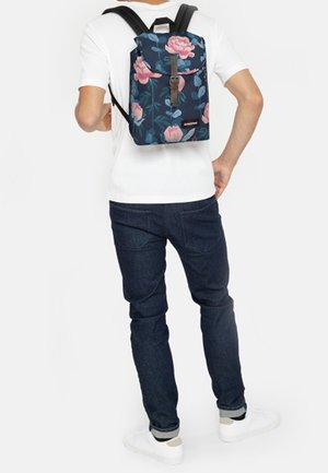 Homme portant un jean foncé et une chemise blanche avec un sac à dos bleu marine présentant un grand motif floral rose et une sangle à boucle en cuir.