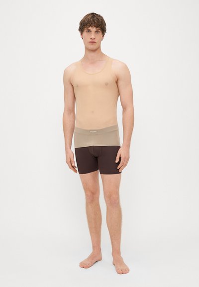 Bēša bez piedurknēm apakšveļa, izgatavota no viegla materiāla, saskaņota ar tumši brūnām pieguļošām šortiem. Vienkāršs dizains, bezšuvju konstrukcija.