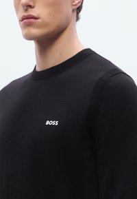 Musta pyöreäkauluksinen neulepaita, joka on valmistettu neuloksesta. Siinä on valkoinen "BOSS"-logo rinnassa sekä resorit kauluksessa ja olkapäällä.