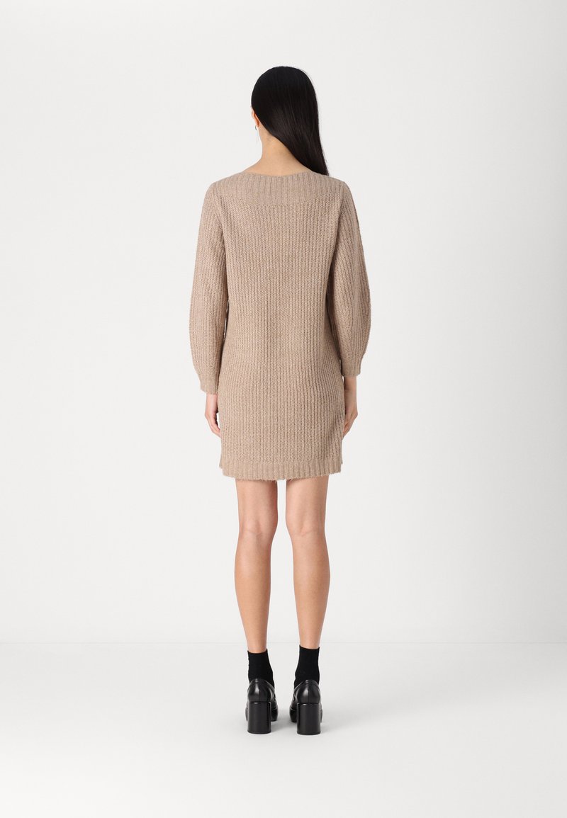 Strickkleid in Hellbeige mit geripptem Muster, Ballonärmeln und einem runden Rückenausschnitt, mit einem leicht ausgestellten Saum. Es werden schwarze Plateauschuhe getragen.