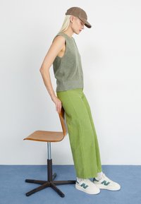 Helles grünes ärmelloses Strickoberteil mit lockerer Passform, kombiniert mit weit geschnittenen, leuchtend grünen Cordhosen. Das Model trägt eine braune Mütze und Sneakers.