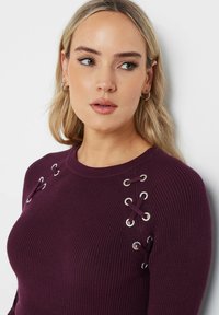 Bordeaux ribbeltrui met metalen oogjes en vetersluiting op de schouders, slanke pasvorm, ronde halslijn en zachte textuur.
