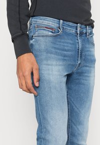 Vaqueros de mezclilla azul ajustados con un lavado desgastado, cinco bolsillos y sutiles detalles de costura roja. Tejido de algodón con una textura suave.