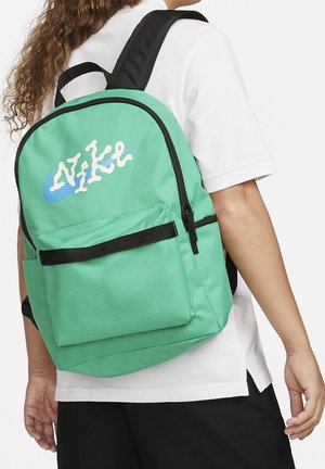 Person iført en grøn Nike-rygsæk med sorte remme og en frontlomme, iført en hvid skjorte og sorte shorts.