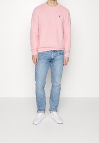 Pull rose clair à col rond avec un petit logo bleu sur la poitrine, associé à un jean bleu clair et des baskets blanches.