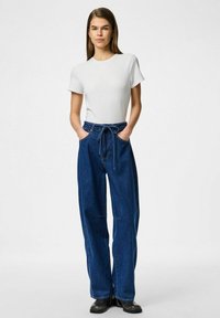 Bjela majica u kombinaciji s plavim visokim širokim trapericama. Traperice imaju pojas s vezicom i dva bočna džepa. Crne čizme upotpunjuju izgled.