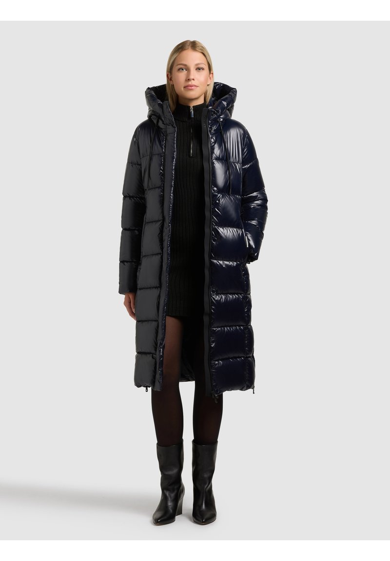 Gara, pūkaina tumši zila jaka ar spīdīgu, izšūtu dizainu, rāvējslēdzēja aizdari un kapuci. Valkāta virs melnas džemperkleitas un zeķbiksēm.