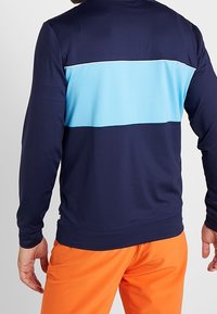 Námořnický modrý top s dlouhým rukávem s světle modrým horizontálním pruhem na zádech, vyrobený z hladkého, elastického materiálu. Viditelné oranžové kalhoty.