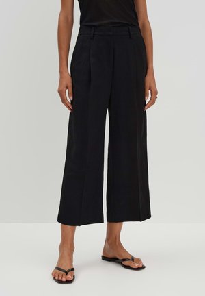 Kobieta ubrana w czarne spodnie z szerokimi nogawkami o wysokim stanie, sięgające do połowy łydki, oraz czarne, otwarte sandały na płaskim obcasie, stojąca na jasnym, jednolitym tle.