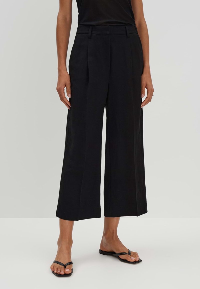 Femme portant un pantalon large noir taille haute et raccourci, et des sandales plates noires à bout ouvert, debout devant un fond clair uni.