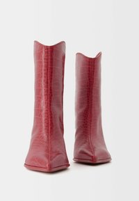 Red ankle boots made of glossy crocodile-patterned material. Features a pointed toe and a distinctive scalloped top edge. Smooth texture.