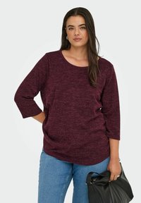 Top bordeaux con texture, maniche a tre quarti, scollatura rotonda e vestibilità ampia, abbinato a jeans blu chiaro e una borsa nera.
