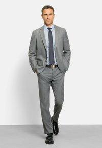 Grauer Nadelstreifenanzug mit einem hellblauen Hemd und einer dunklen Krawatte. Hat einen maßgeschneiderten Schnitt und elegante schwarze Schuhe. Das Model steht selbstbewusst mit den Händen in den Taschen.