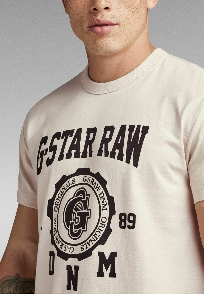Jeune personne avec des taches de rousseur portant un t-shirt beige clair avec un grand texte noir "G-STAR RAW" et un logo circulaire avec les lettres "89" et "DNM".
