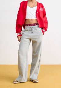 Osoba ubrana w krótką czerwoną kurtkę, białą bluzkę crop top, szare dresy Nike, beżowe sneakersy i widoczną bordową gumę od spodenek Nike, stojąca na żółtej podłodze.