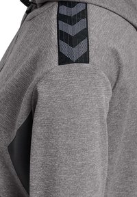 Grijze sportieve hoodie met een textuur, zwarte panelen en een kenmerkend chevron patroon op de schouder in zwart en wit.