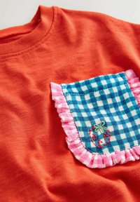 Lyseorange stof med en blå og hvidternet lomme kantet med lyserøde flæser og broderet med en lille rød jordbær.