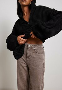 Schwarzer gerippter Pullover mit hohem Kragen und Reißverschluss vorne, kombiniert mit beigen Jeans mit subtiler Ausbleichung und klassischem Fünf-Taschen-Design.
