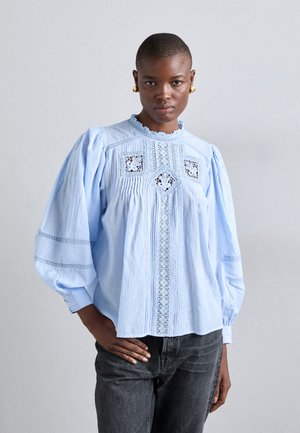 Világoskék blúz, tágas ujjal, hímzett részletekkel és csipkés díszítéssel az elején és az ujjak végén. Sötétszürke farmernadrággal párosítva.