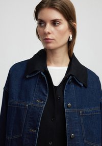 Chaqueta de denim con un cuello negro en contraste, tela de color azul oscuro, bolsillos frontales y herrajes plateados. Lleva encima una camisa negra.