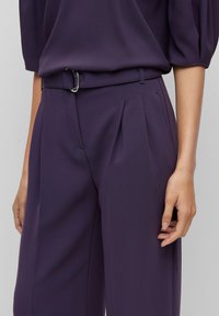 Kalhoty s širokými nohavicemi v tmavě fialovém materiálu, se sklady vpředu, páskem se stříbrnou sponou a hladkým povrchem. Ruka umístěná na pase.