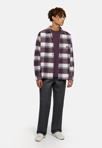 Lila och vit rutig skjorta över en mörklila t-shirt, i kombination med grå byxor och vita sneakers. Skjortan har knäppning och ficka.