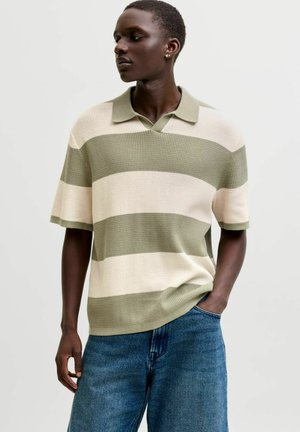Personne aux cheveux courts portant un polo en maille à manches courtes rayé vert et beige et un jean bleu, debout avec une main dans la poche.
