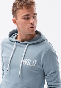 Felpa leggera blu con cappuccio, con una tasca a marsupio frontale, cordini bianchi e una stampa sbiadita "WILD" sul petto in un carattere usurato.