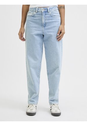 Personne portant un jean bleu clair taille haute, jambes droites, et des baskets blanches, debout devant un fond blanc uni.