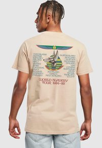 Beige bomull t-skjorte med grafikk av et stilisert hundehode med vinger, og tekst som oppgir land knyttet til en turné i 1984-85.