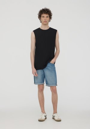 Maglietta nera senza maniche abbinata a pantaloni shorts in denim arrotolati. L'outfit è completato da sneakers bianche con accenti neri.