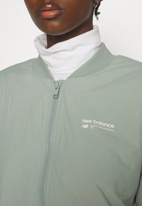 Veste bomber vert clair avec une fermeture éclair à l'avant, un col côtelé et un logo blanc imprimé sur la poitrine. Superposée sur un col roulé blanc.