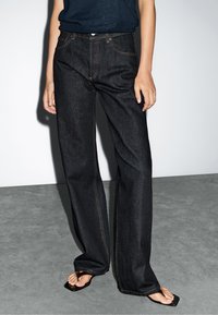 Jeans a gamba larga in denim blu scuro con due tasche frontali, cuciture arancioni e un classico design a cinque tasche, abbinati a infradito nere.