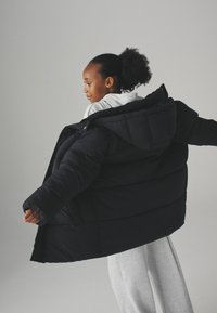 Sort puffjakke med hætte, der har et quiltet design og løs pasform. Båret med lysegrå teksturerede bukser.