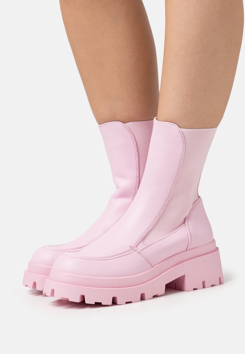 Jambes portant des bottes roses claires mi-mollet, à semelle épaisse et panneaux latéraux élastiques sur fond blanc.