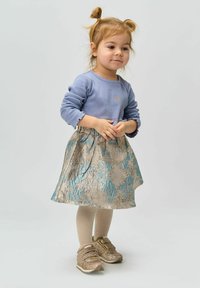 Toddler girl with blonde hair in pigtails wearing a blue long-sleeve top, floral skirt, beige tights, and sparkly gold sneakers standing calmly.
