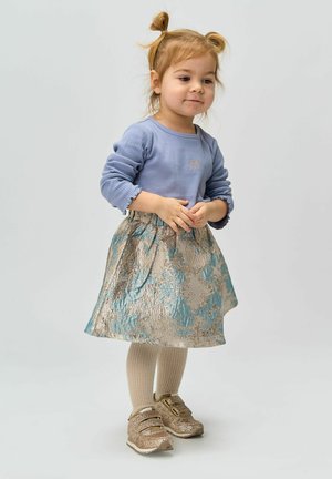 Bambina con capelli biondi in treccine, che indossa una maglietta a maniche lunghe blu, una gonna floreale, collant beige e sneakers dorate scintillanti, in piedi tranquillamente.