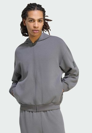 Personne avec des dreadlocks portant un sweat à capuche gris zippé et un pantalon assorti, debout les mains dans les poches sur un fond uni.