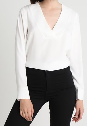 Femme portant un chemisier blanc à manches longues et col en V, rentré dans un pantalon noir taille haute, sur un fond uni.