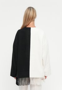 Mulher com cabelo loiro longo usa um suéter oversized em preto e branco com franjas combinando na barra, de pé contra um fundo branco.