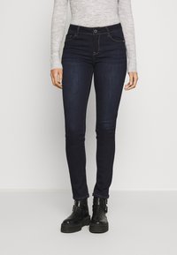 Tmavě modré skinny džíny s hladkým povrchem, designem s pěti kapsami a kontrastním prošíváním. Nosit s černými kotníkovými botami.